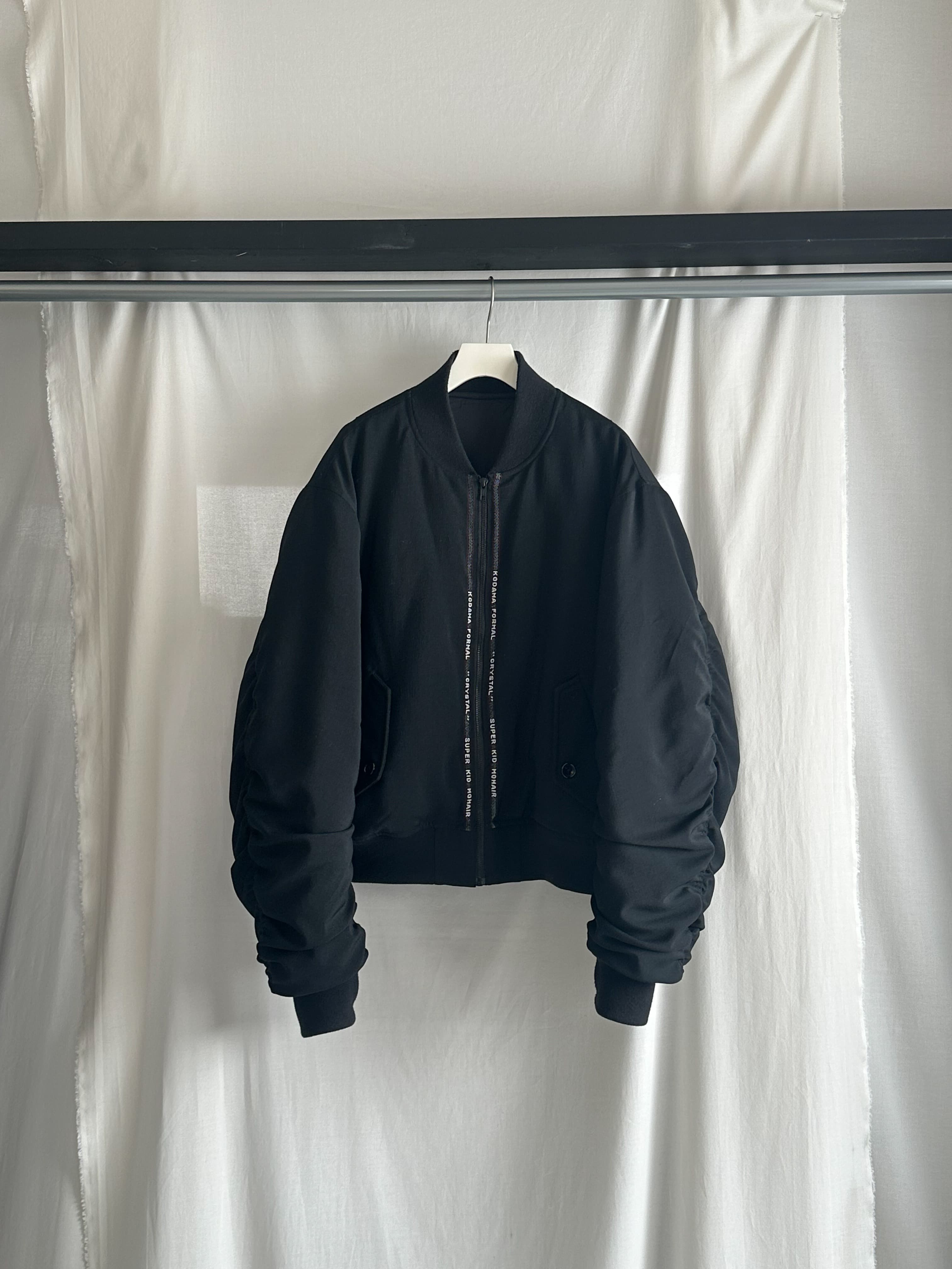 キッドモヘア混の高級スーツ生地をベースに中綿を入れボンバージャケットを制作。前中心を生地耳で装飾し、フラップポケットを採用。厳つさと高級感が合わさった一着。